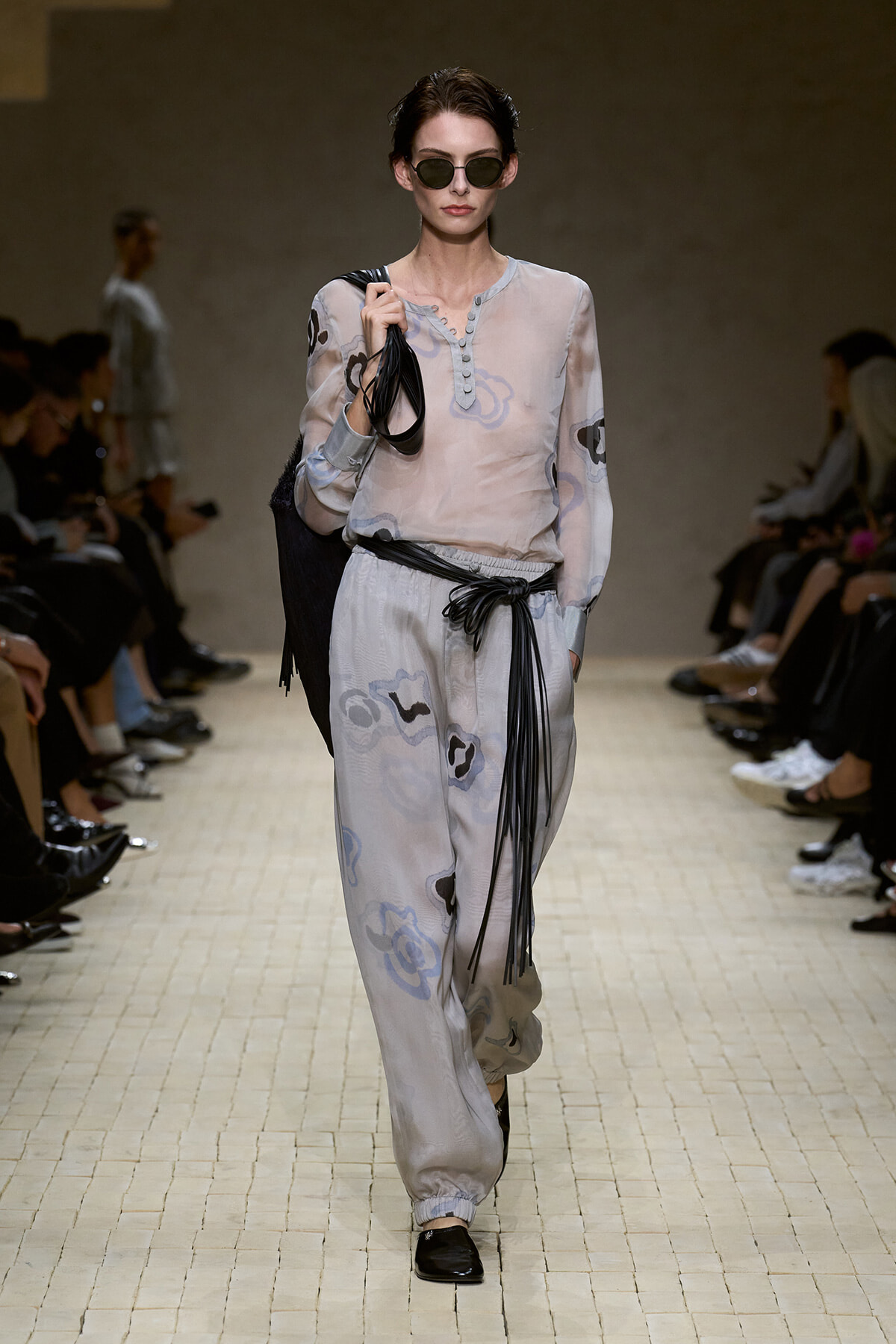 Model walks a fashion runway in a pale floral-pattern blouse and matching wide-leg pants, with a black fringe belt and sunglasses, carrying a black bag on her shoulder.