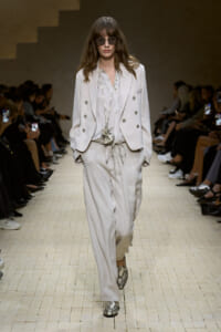 Model walks the runway in a light gray oversized double-breasted suit with wide-leg pants and snake-print loafers, wearing round sunglasses and a long silver necklace.