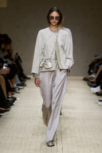 Runway model in a pale gray textured cardigan with matching top and wide-leg satin pants, accessorized with two small crossbody bags and sunglasses.
