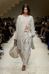 Runway model in a loose cream coat over pale pants, with a large textured beige tote bag and neutral flats.