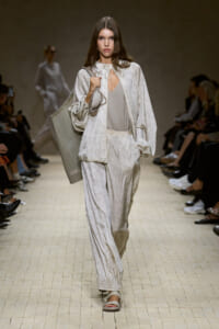 Model walks a runway in a pale gray velvety matching suit with wide-leg pants, carrying a large pleated gray tote on her shoulder