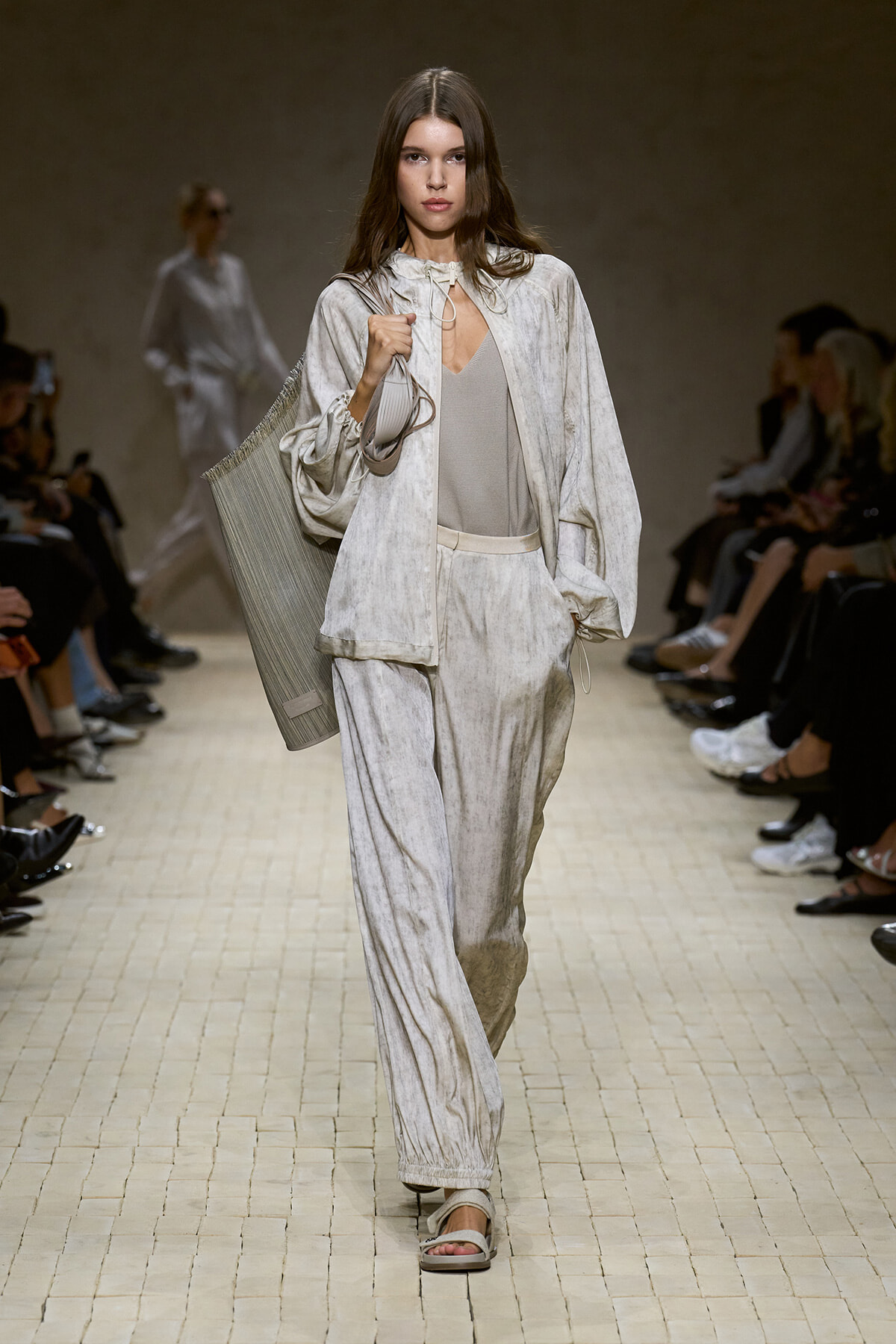Model walks a runway in a pale gray velvety matching suit with wide-leg pants, carrying a large pleated gray tote on her shoulder