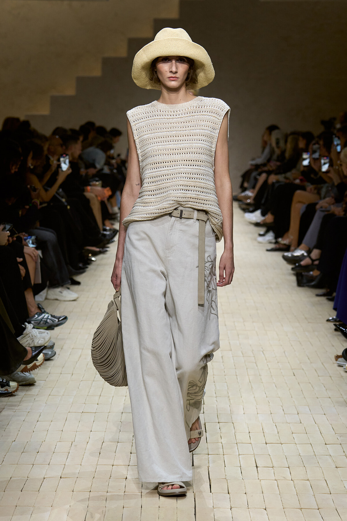 Model walking a fashion runway in a beige knitted sleeveless top, wide-leg light gray pants with a belt, beige sandals, and a large straw hat while carrying a pleated beige bag.