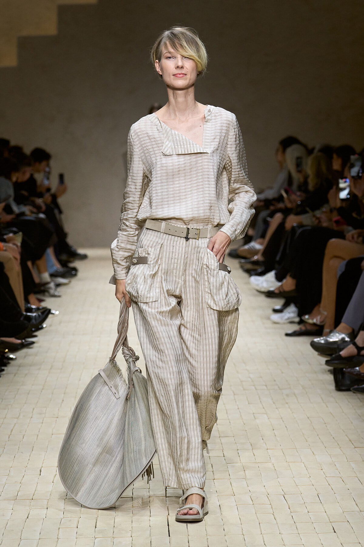 Model walking runway in beige striped outfit with belt, carrying large gray striped tote bag.