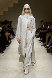 Model walks a light beige patterned coat over wide-leg satin pants on a runway, wearing sunglasses.