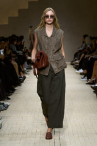 Fashion model walks a runway in a textured brown sleeveless vest over olive wide-leg pants, carrying a burgundy bag and wearing rounded sunglasses.