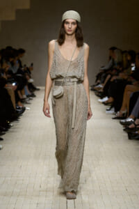 Model walking a beige patterned jumpsuit with a matching belt, small crossbody bag, and a textured cream headscarf on a fashion show runway