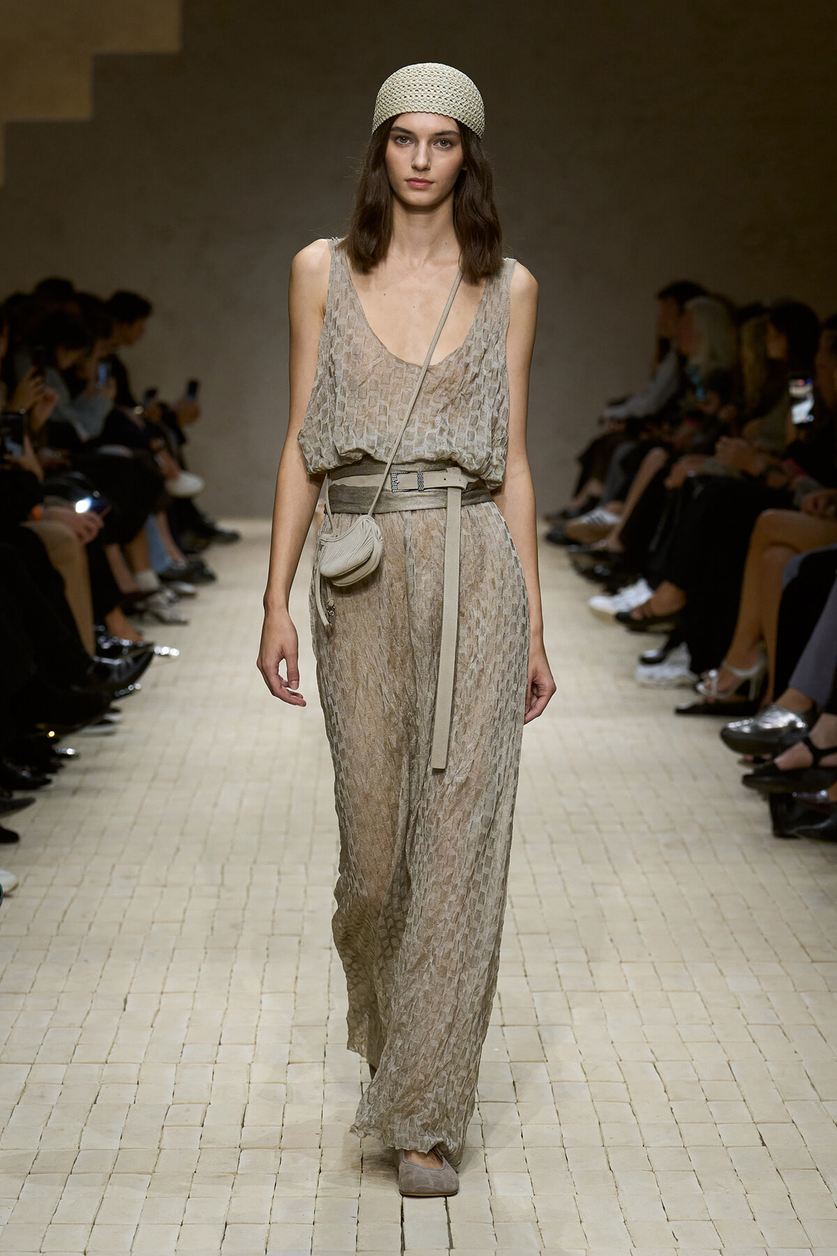Model walking a beige patterned jumpsuit with a matching belt, small crossbody bag, and a textured cream headscarf on a fashion show runway
