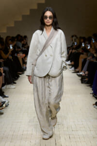 Model on a runway wearing an oversized cream blazer, wide leg patterned pants, and a beige crossbody strap bag with dark sunglasses.