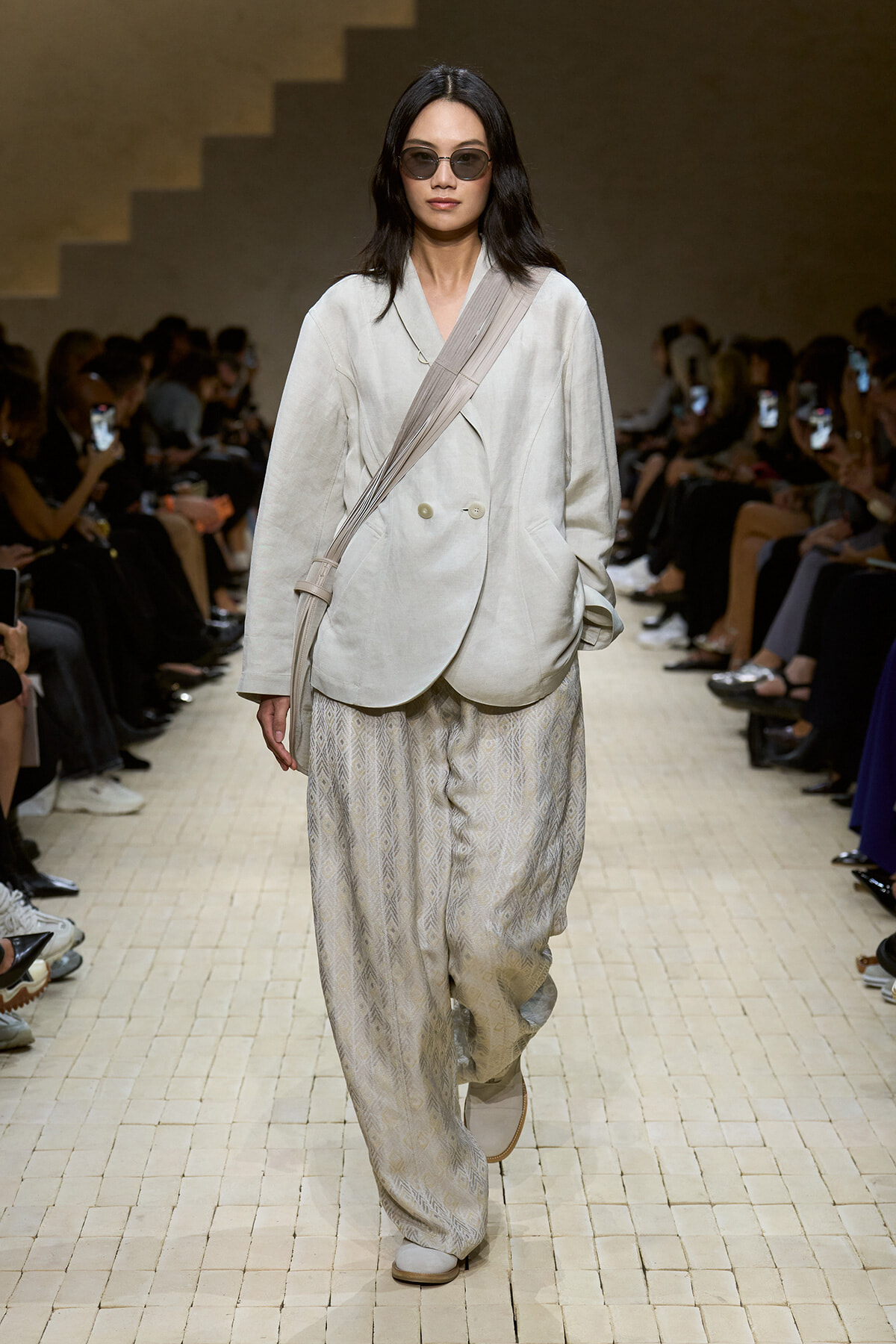Model on a runway wearing an oversized cream blazer, wide leg patterned pants, and a beige crossbody strap bag with dark sunglasses.