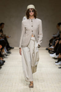 Model walks the runway in a light beige striped jacket with large buttons, paired with wide, braided-strap linen pants and a woven belt fringe detail.