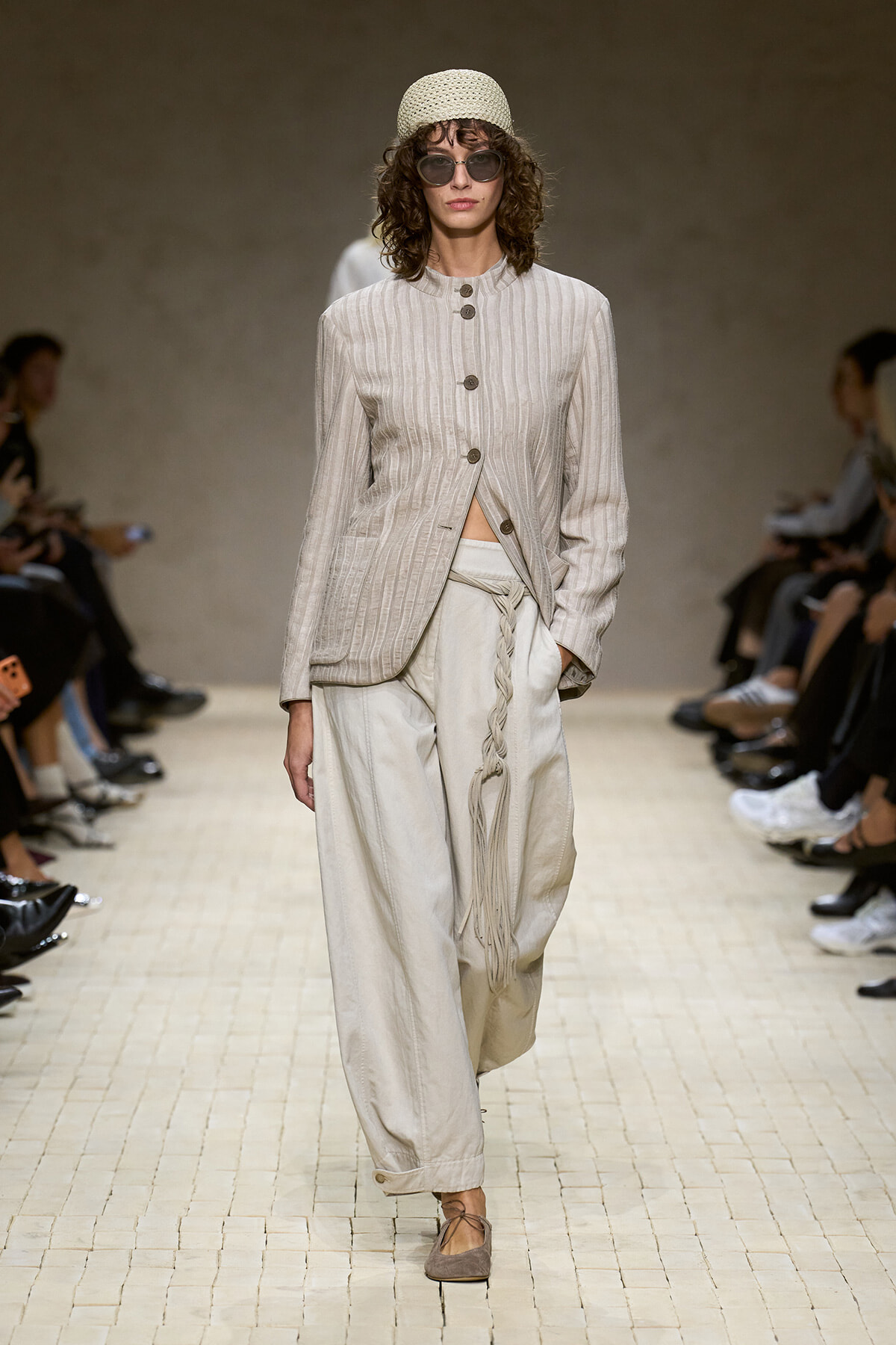 Model walks the runway in a light beige striped jacket with large buttons, paired with wide, braided-strap linen pants and a woven belt fringe detail.