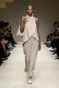 Model on a runway wearing a beige draped sleeveless top, striped wide-leg pants, white sneakers, and a woven beige tote bag