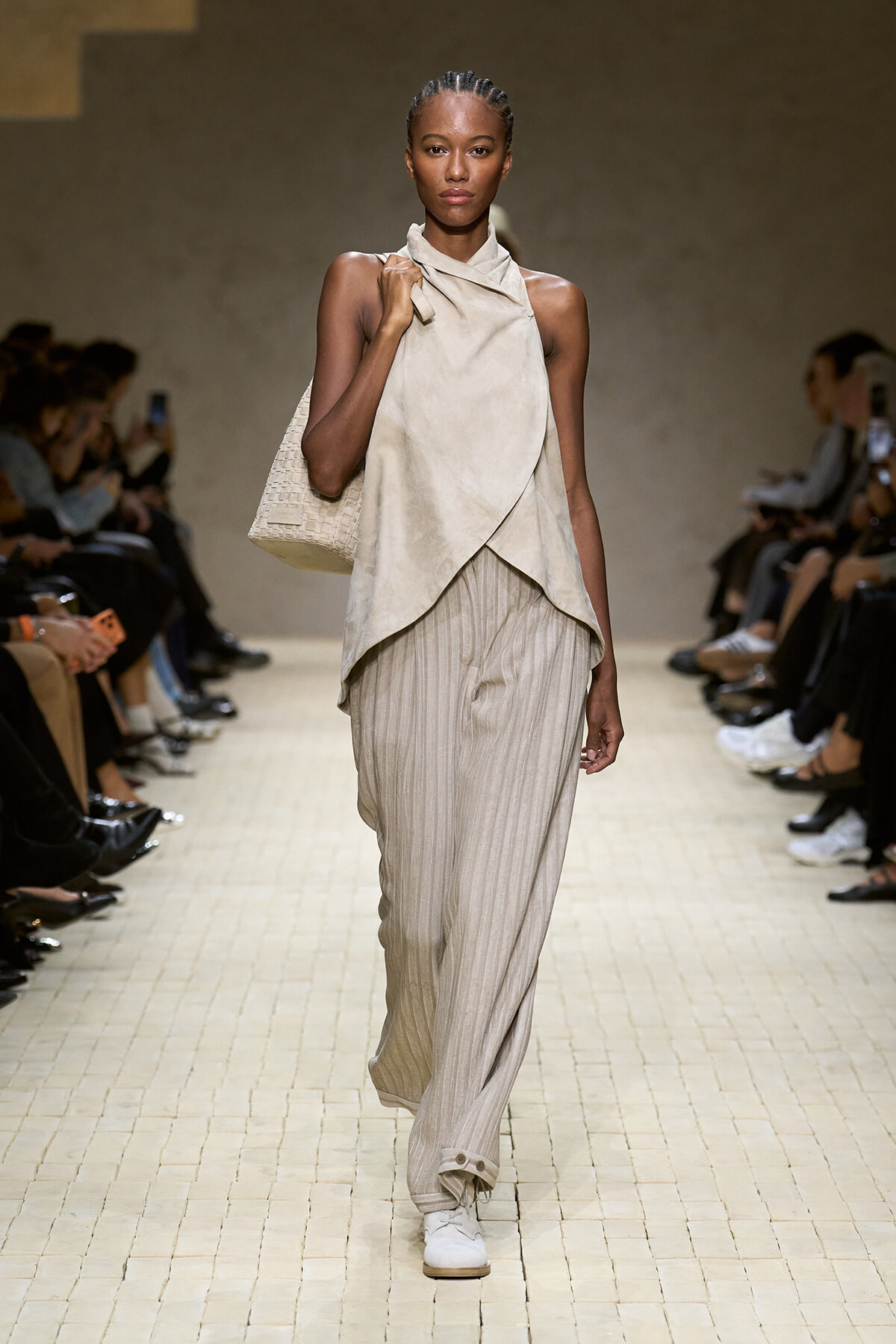 Model on a runway wearing a beige draped sleeveless top, striped wide-leg pants, white sneakers, and a woven beige tote bag