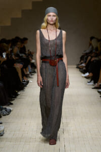 Model on a runway wearing a sheer gray maxi dress, a red belt with a small round brown pouch, and a gray knit headband.