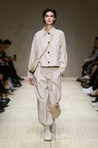 Model on a runway wearing a beige oversized jacket with matching wide-leg pants, crossbody bag, and light boots.