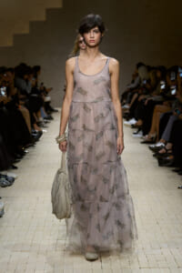 Model walks the runway in a sheer, dusty-pink, tiered maxi dress with subtle floral prints, carrying a beige handbag.
