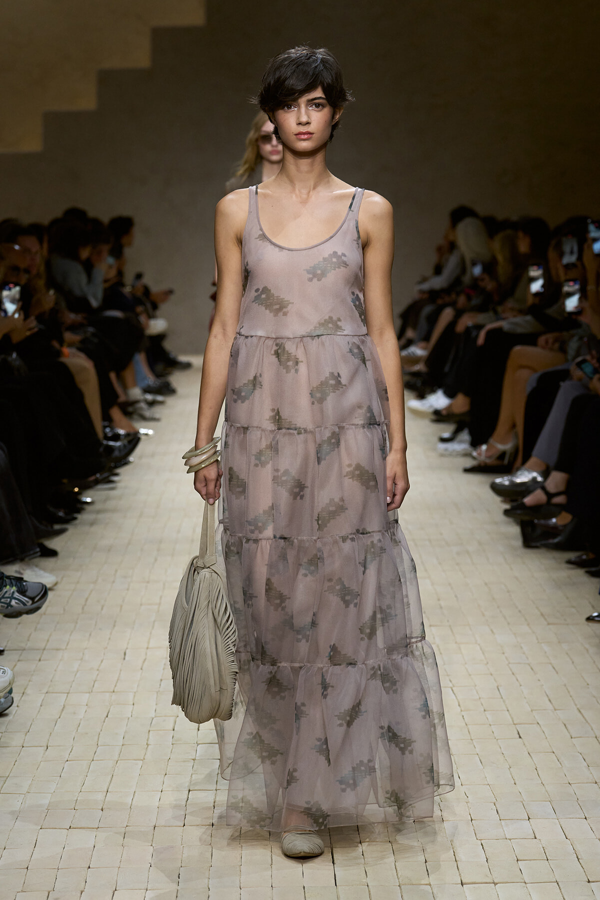 Model walks the runway in a sheer, dusty-pink, tiered maxi dress with subtle floral prints, carrying a beige handbag.