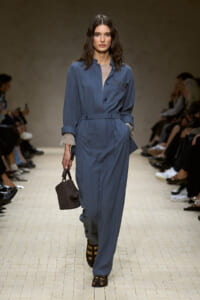 Model walking the runway in a blue jumpsuit with belt, rolled cuffs, and a small brown woven handbag in hand.