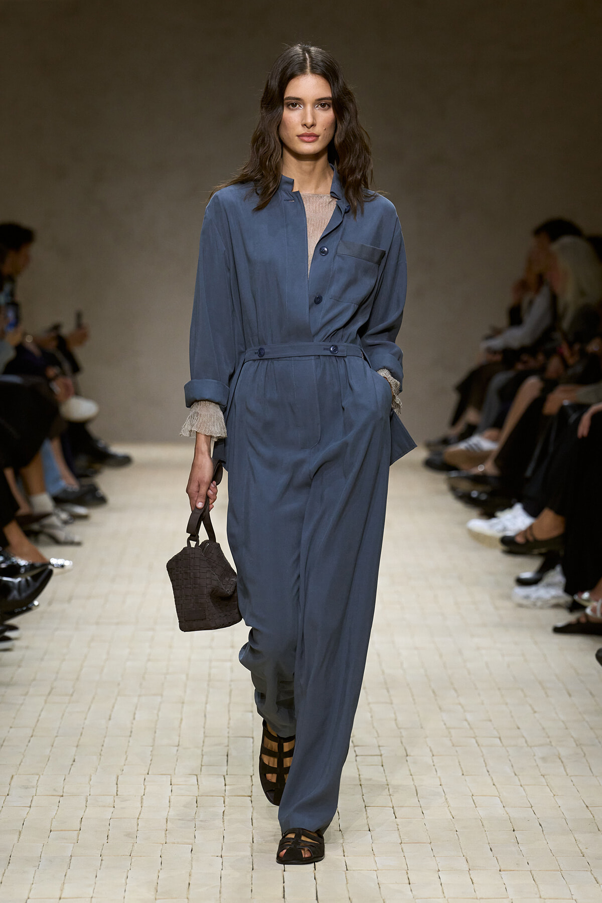 Model walking the runway in a blue jumpsuit with belt, rolled cuffs, and a small brown woven handbag in hand.