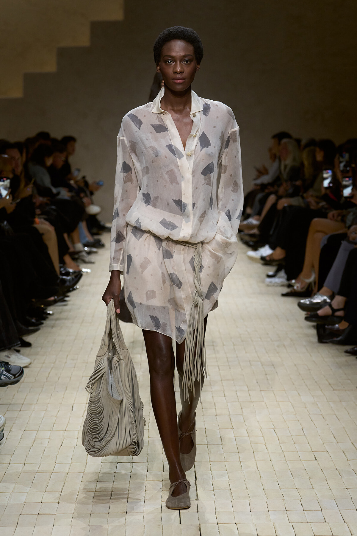 Model walking a fashion runway in a sheer cream blouse with gray abstract prints and matching shorts, carrying a large fringed beige bag.