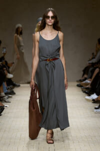 Model walks a fashion runway in a gray sleeveless maxi dress with a tied waist, wearing sunglasses and carrying a large brown tote.
