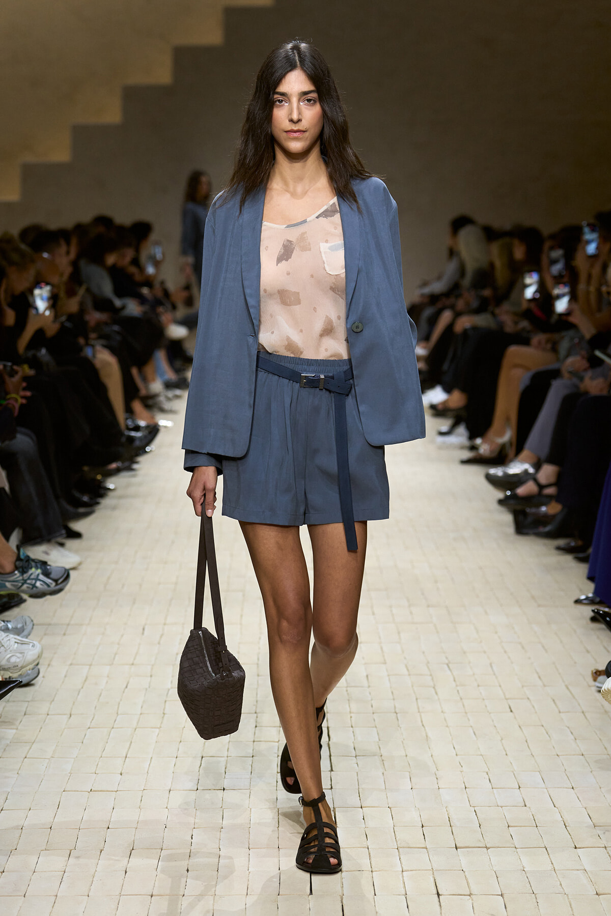 Female model walking a runway in a blue blazer and matching shorts, with a beige patterned top and a small black woven handbag.