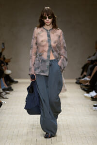 Model walking a runway in a sheer pastel floral cardigan, loose blue-gray pants, dark sunglasses, and carrying a large navy pleated bag with a tied wrist accessory.