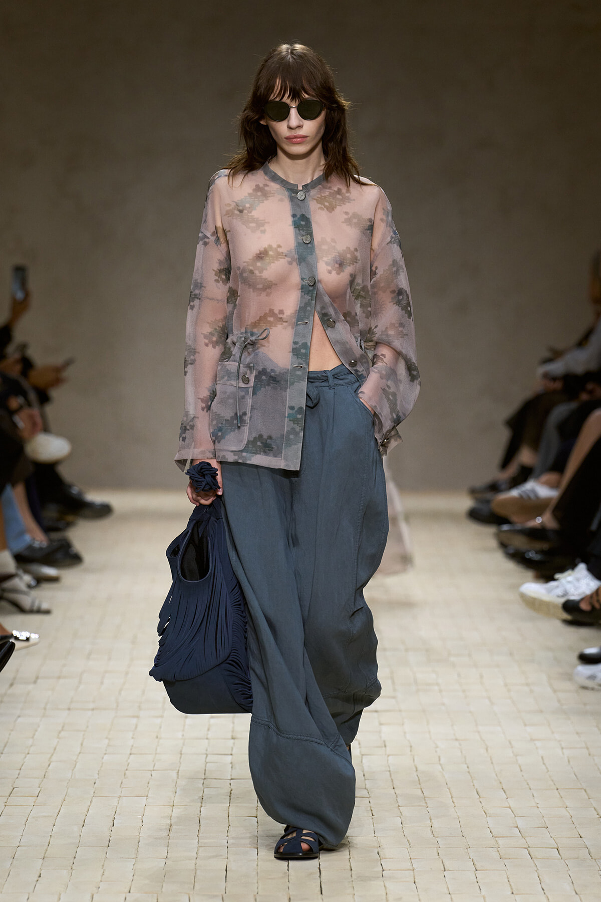 Model walking a runway in a sheer pastel floral cardigan, loose blue-gray pants, dark sunglasses, and carrying a large navy pleated bag with a tied wrist accessory.