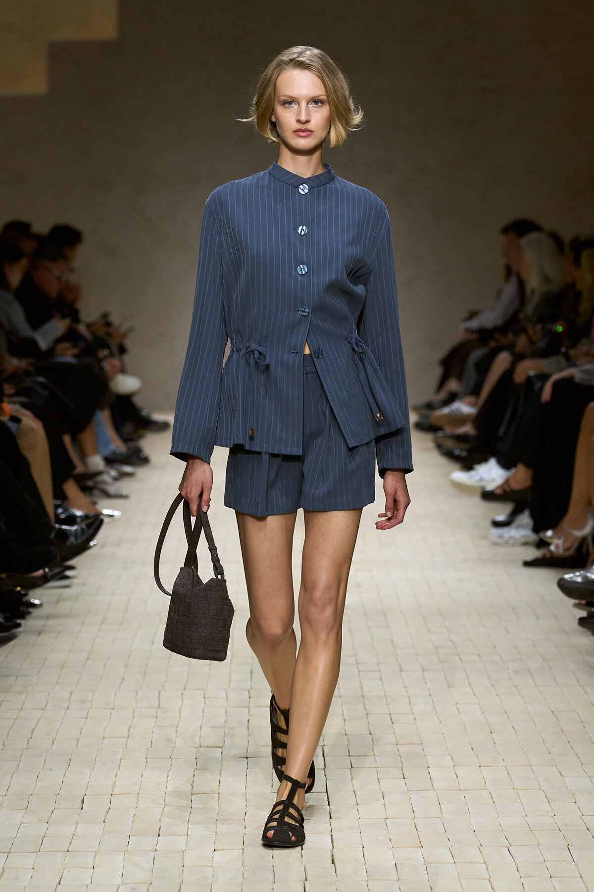 Model walks a fashion show wearing a blue pinstripe matching jacket and shorts, carrying a brown woven handbag.
