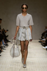 Model on a runway wearing a light gray short-sleeve polo top, a pleated patterned mini skirt, and a beige belt, carrying a large light gray handbag and wearing sunglasses.