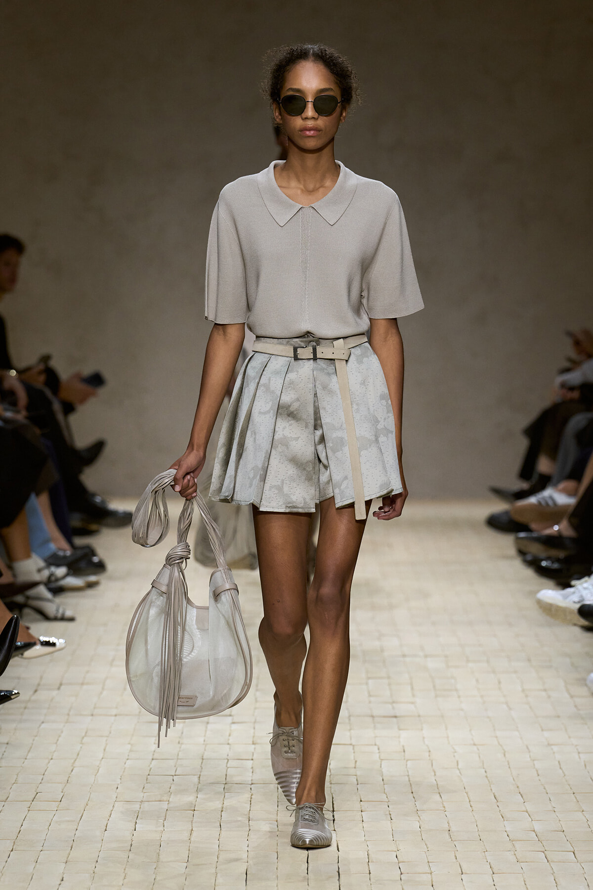 Model on a runway wearing a light gray short-sleeve polo top, a pleated patterned mini skirt, and a beige belt, carrying a large light gray handbag and wearing sunglasses.