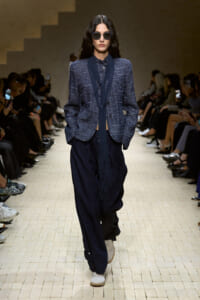 Model on a runway wearing a navy textured blazer with a long scarf, loose dark navy pants, white sneakers, and round sunglasses.