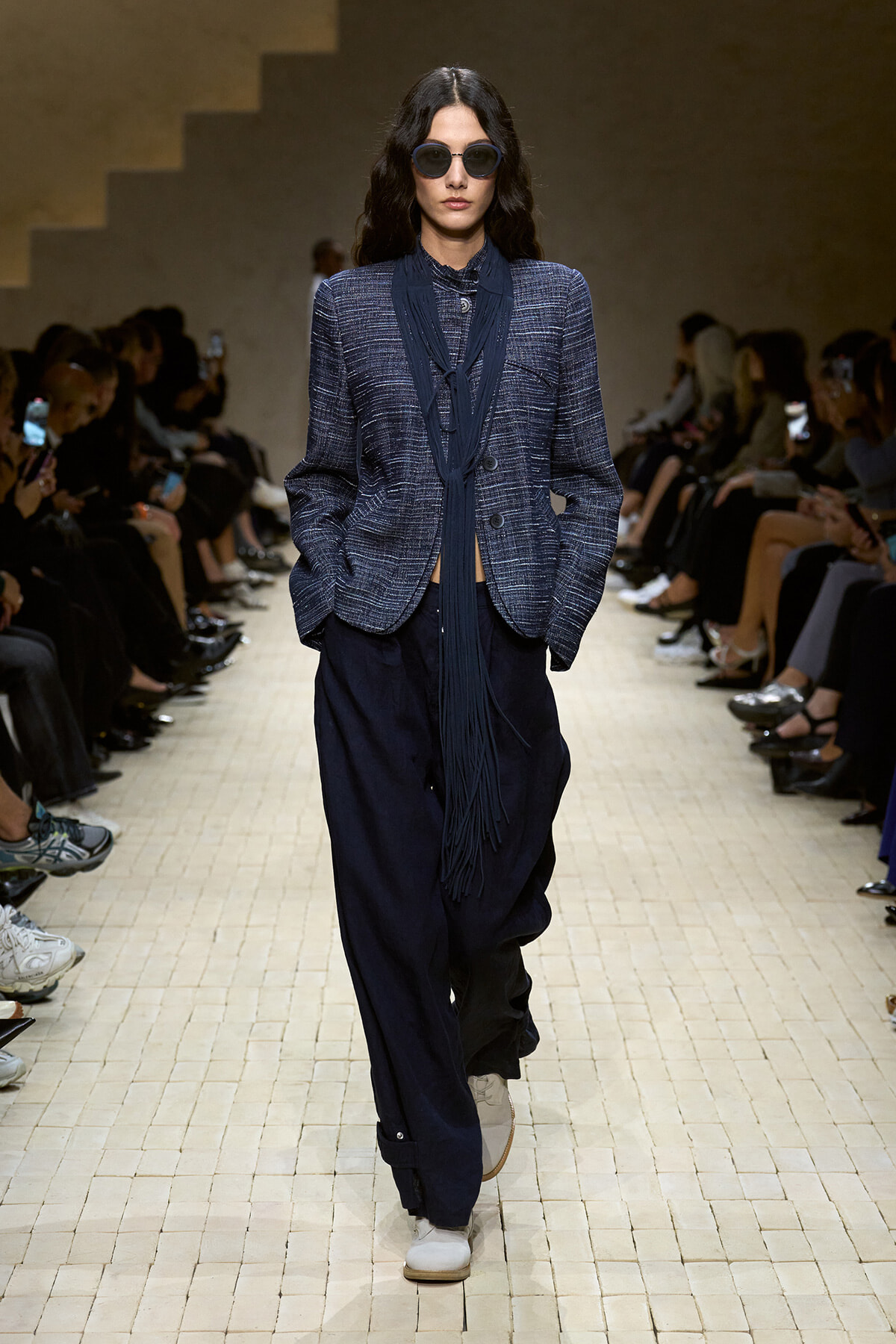 Model on a runway wearing a navy textured blazer with a long scarf, loose dark navy pants, white sneakers, and round sunglasses.