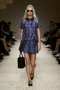 Model on runway wearing a violet patterned blouse with a dark collar and a pleated denim mini skirt, carrying a black handbag and wearing sunglasses.