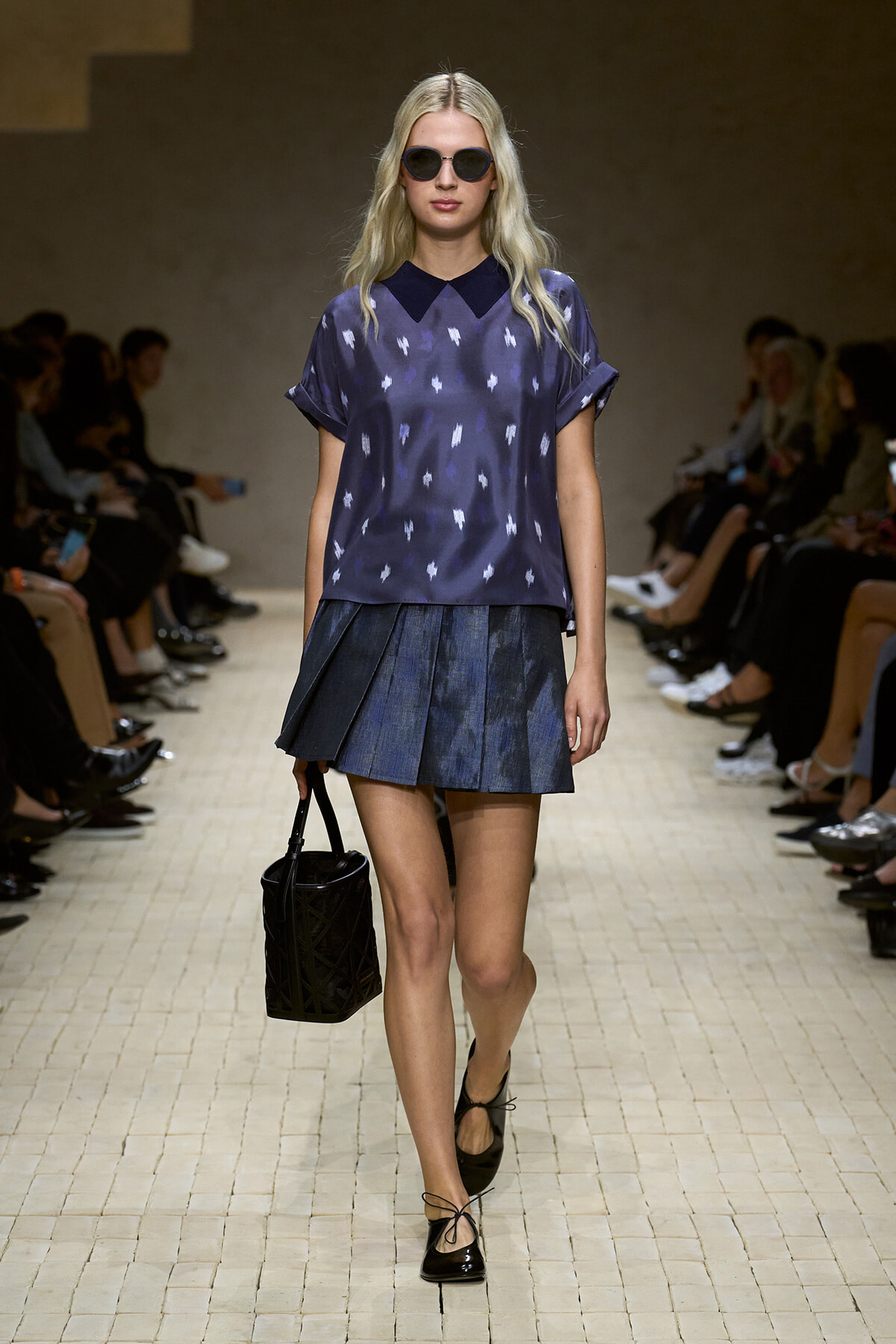Model on runway wearing a violet patterned blouse with a dark collar and a pleated denim mini skirt, carrying a black handbag and wearing sunglasses.