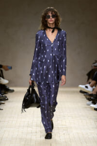 Runway model in a navy blue patterned suit with a black neck scarf, round sunglasses, and a black fringed handbag.