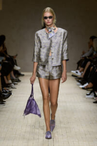 Model on a runway wearing a lavender checkered suit with a matching short blazer and shorts, sunglasses, and a purple handbag.