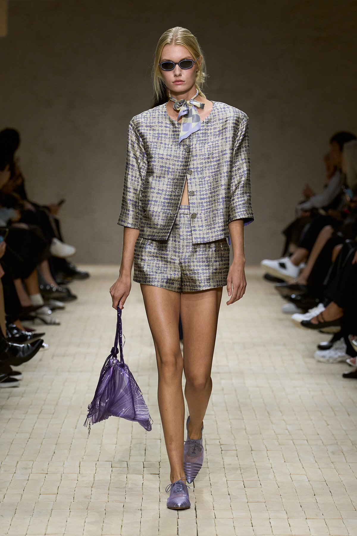 Model on a runway wearing a lavender checkered suit with a matching short blazer and shorts, sunglasses, and a purple handbag.