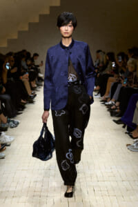 Model walking a fashion runway in a cobalt blue satin jacket, black printed pants, and a dark blue handbag.