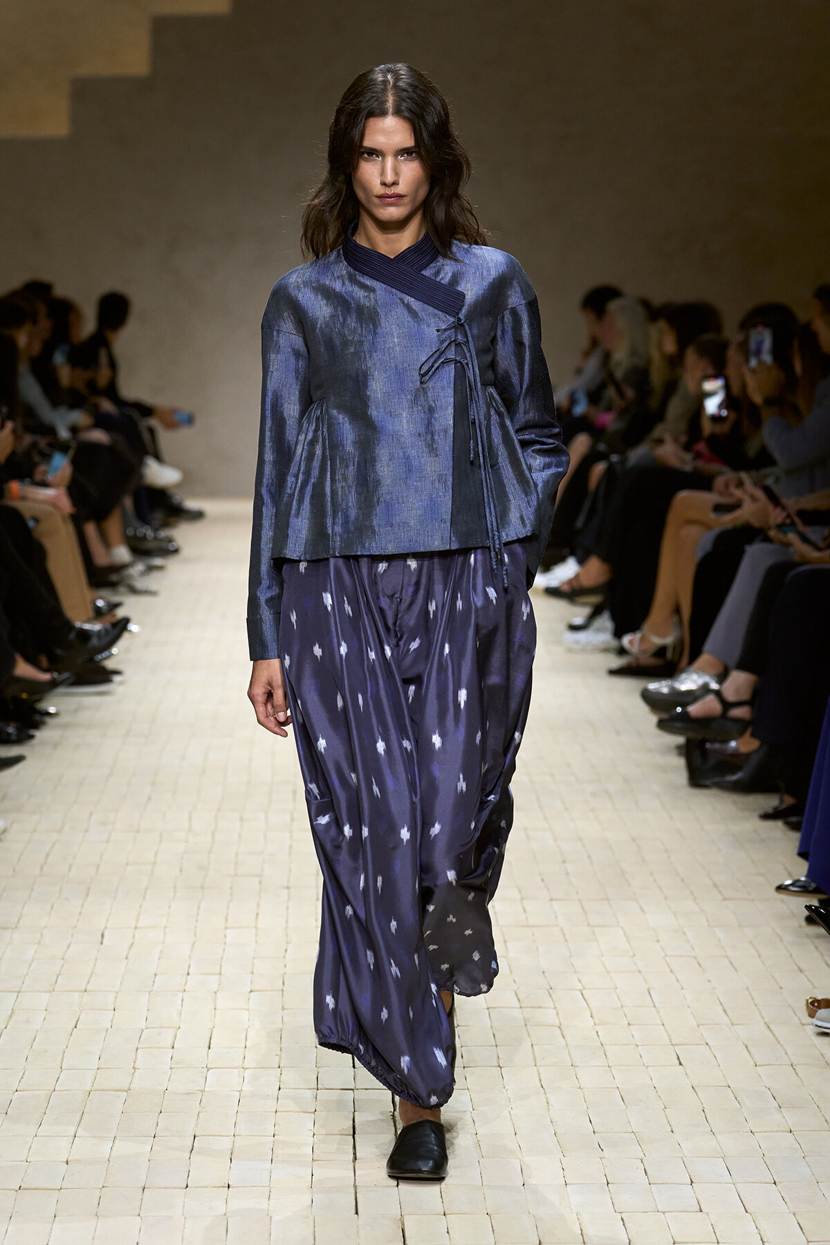 Model walking runway in a blue, textured wrap-top and loose cobalt pants, audience on both sides observing Creased light-colored tiles runway.