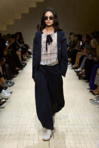 Model walks a runway wearing a navy coordinating suit with wide-leg pants and a sheer checkered blouse tied with a black bow, sunglasses on.