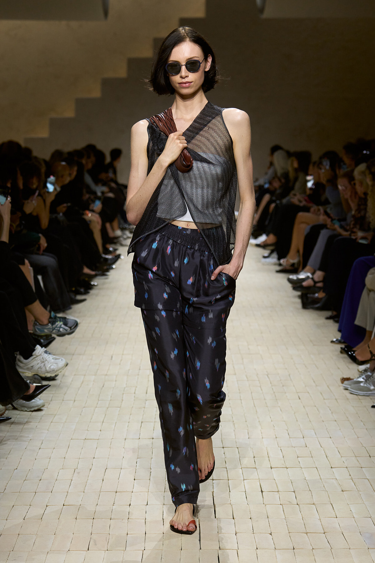 Model walks a fashion show runway in a sheer black mesh layered top, dark patterned pants, and sunglasses, carrying a brown bag strap over her shoulder.