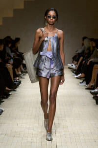 Model walking a runway in a metallic gray sleeveless top and matching shorts with a lavender belt, small sunglasses, and a striped tote over her shoulder.