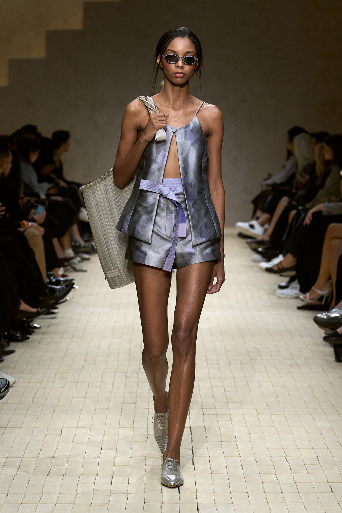 Model walking a runway in a metallic gray sleeveless top and matching shorts with a lavender belt, small sunglasses, and a striped tote over her shoulder.