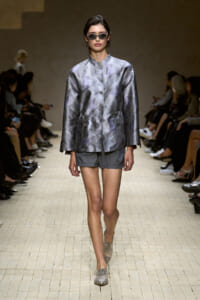 Model on a runway wearing a metallic silver-gray satin jacket with a stand collar, front buttons, drawstring waist, and matching gray shorts, plus sunglasses.