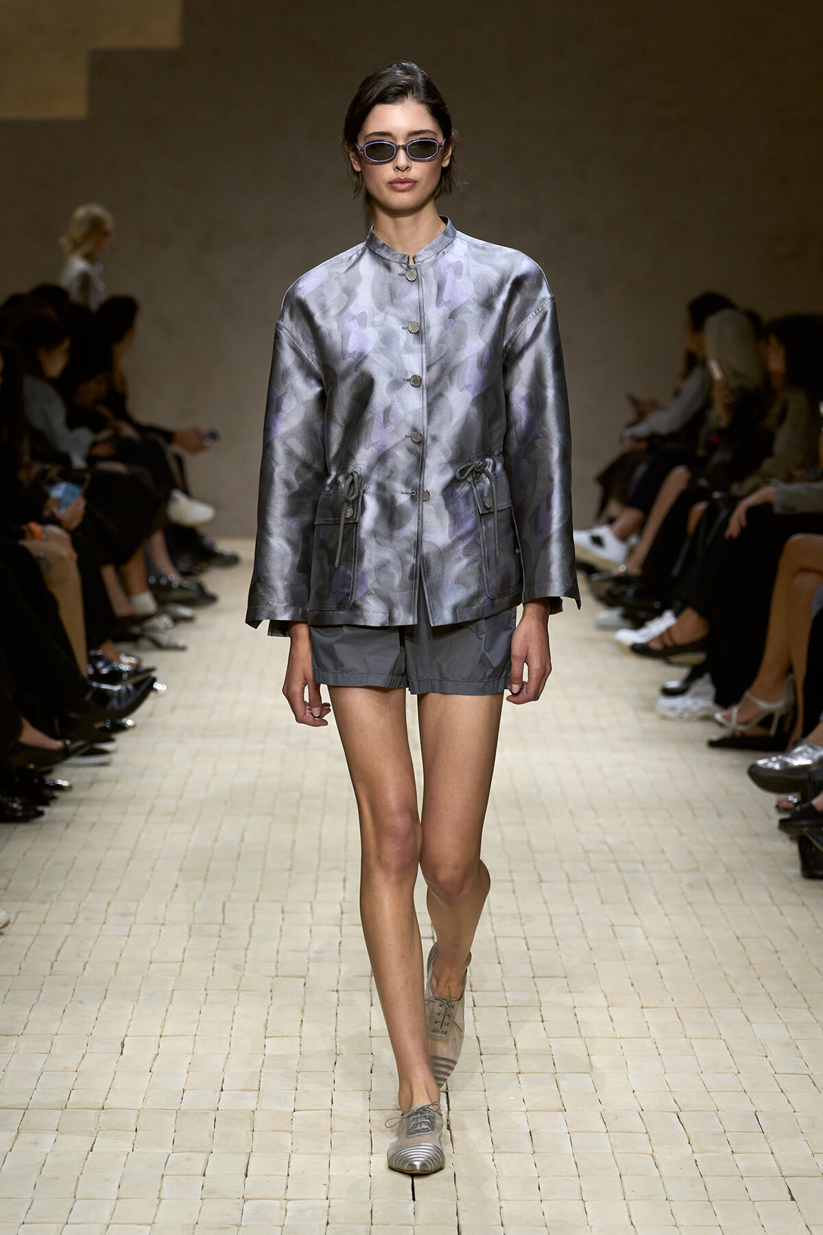 Model on a runway wearing a metallic silver-gray satin jacket with a stand collar, front buttons, drawstring waist, and matching gray shorts, plus sunglasses.