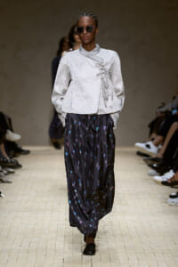 Model walking a runway in a light gray wrap-top with lace details and dark, speckled wide-legged pants; she wears round sunglasses and black shoes.