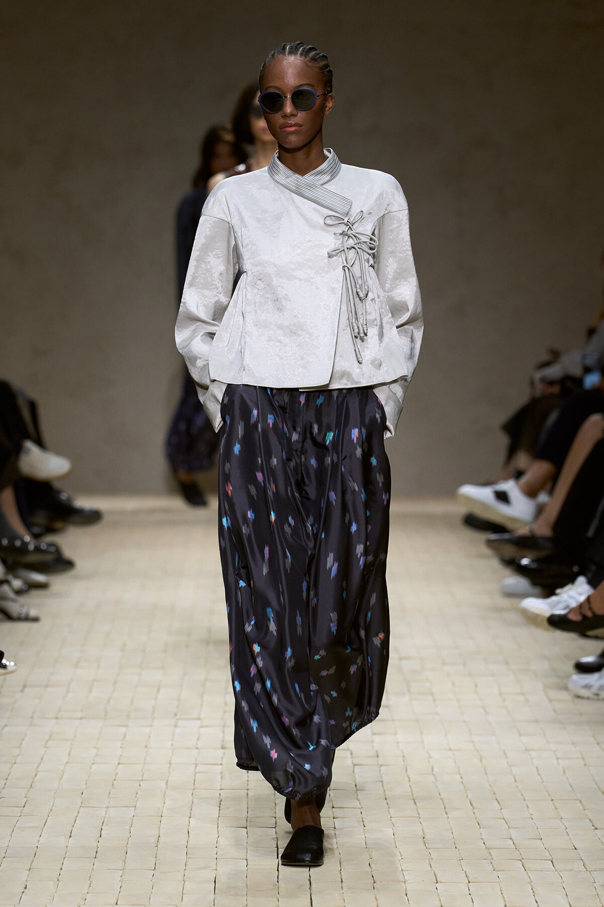 Model walking a runway in a light gray wrap-top with lace details and dark, speckled wide-legged pants; she wears round sunglasses and black shoes.