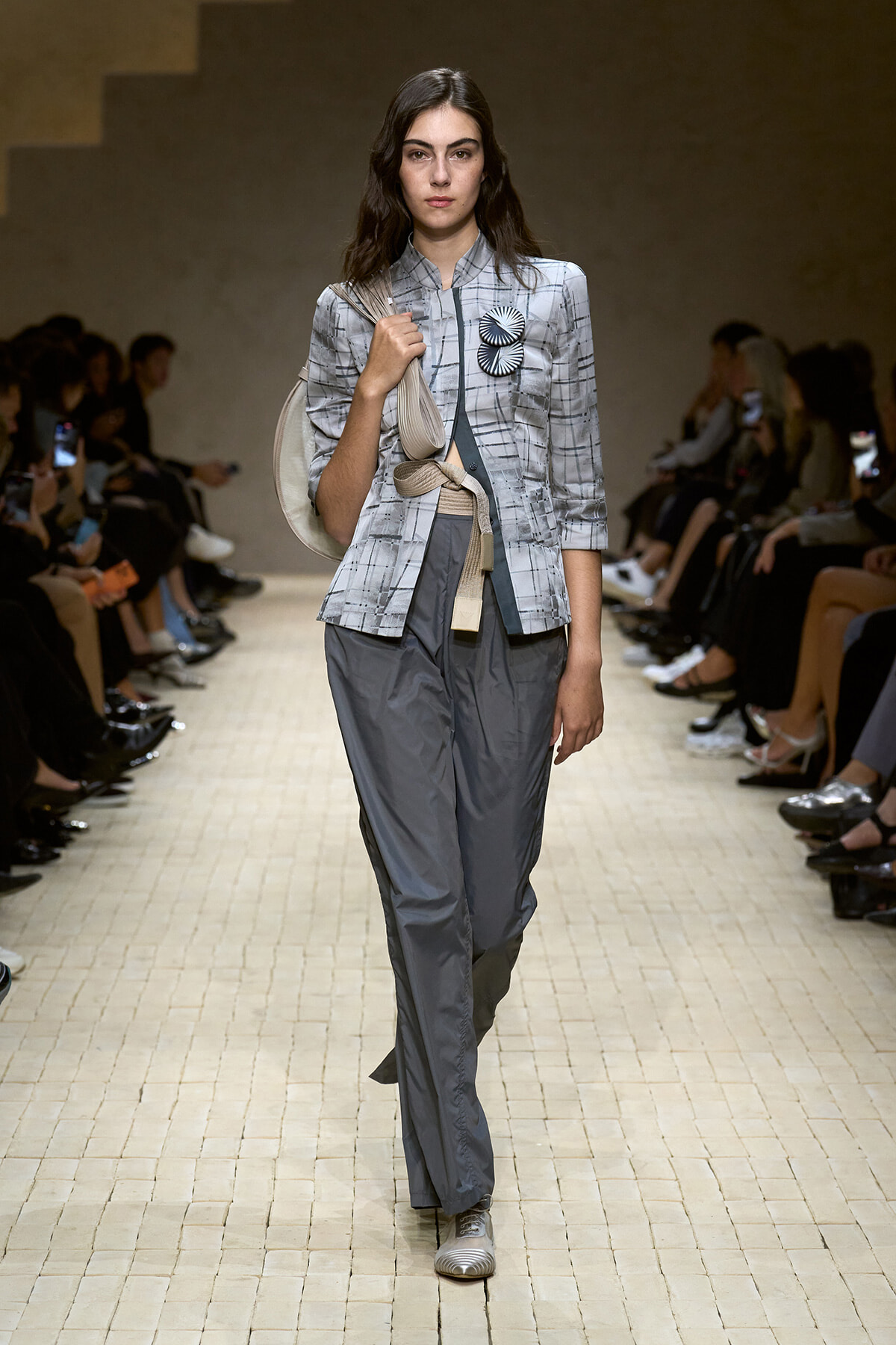 Female model walks a runway wearing a light gray plaid blazer with two round black-and-white badges, gray wide-leg pants, silver shoes, and a beige shoulder bag.